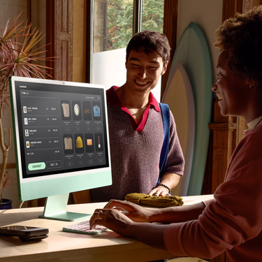 サーフボードなどを販売しているショップのカウンター内に座っている従業員がiMacを操作し、カウンターの反対側にいるお客様の決済処理を行なっている。