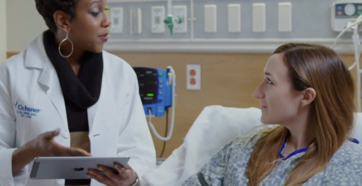 Une soignante tenant un iPad discute avec une patiente installée dans un lit d’hôpital.