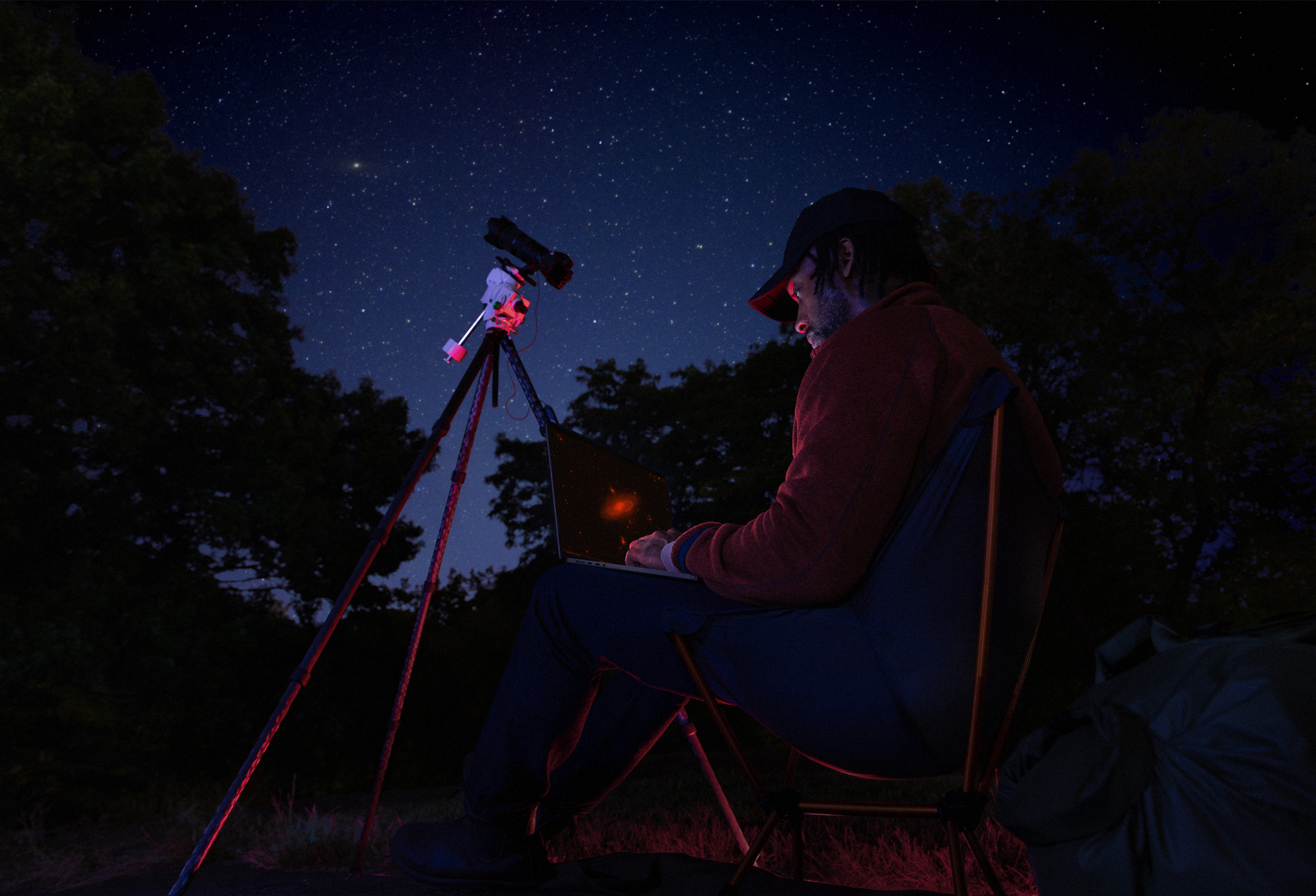A person outside at night using a MacBook Pro on their lap, connected to a device on a tripod pointed toward the starry sky