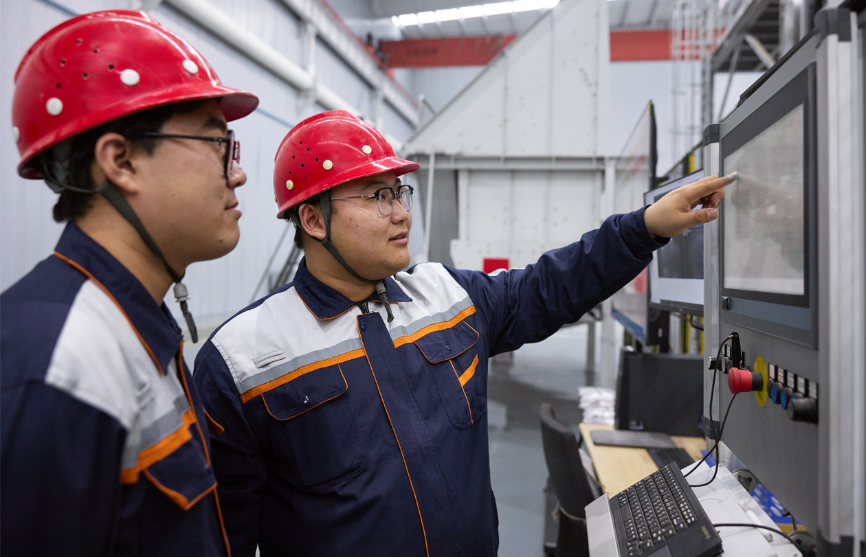 Two employees wearing safety hats and uniforms look at digital screen with control handles at the bottom